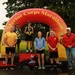 A podium ceremony at the Marine Corps Marathon featuring three place finishers standing on 1st, 2nd, and 3rd place platforms beneath an inflatable arch. The event appears to be taking place in wet, overcast conditions on a paved surface surrounded by green grass and trees. Several officials or staff members flank the podium alongside the medal-wearing competitors.