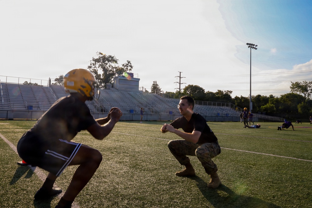 Two individuals face each other in a crouched athletic stance on a green turf football field, appearing to engage in a training or coaching drill. One person wears a gold football helmet and dark athletic shorts, while the other is dressed in camouflage pants and a black t-shirt, suggesting a military or ROTC involvement. Additional players and individuals are visible in the background near stadium bleachers.