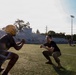 Two individuals face each other in a crouched athletic stance on a green turf football field, appearing to engage in a training or coaching drill. One person wears a gold football helmet and dark athletic shorts, while the other is dressed in camouflage pants and a black t-shirt, suggesting a military or ROTC involvement. Additional players and individuals are visible in the background near stadium bleachers.