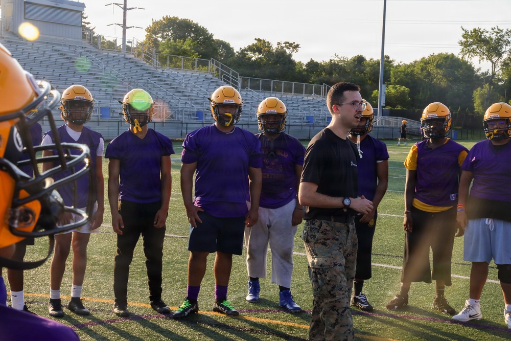A football team practice session is taking place on an outdoor turf field, with players wearing purple jerseys and gold helmets gathered in a group. A man in a black shirt and military camouflage pants, likely a coach or military liaison, appears to be addressing the team. The setting includes stadium bleachers and trees visible in the background.