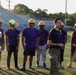 A football team practice session is taking place on an outdoor turf field, with players wearing purple jerseys and gold helmets gathered in a group. A man in a black shirt and military camouflage pants, likely a coach or military liaison, appears to be addressing the team. The setting includes stadium bleachers and trees visible in the background.