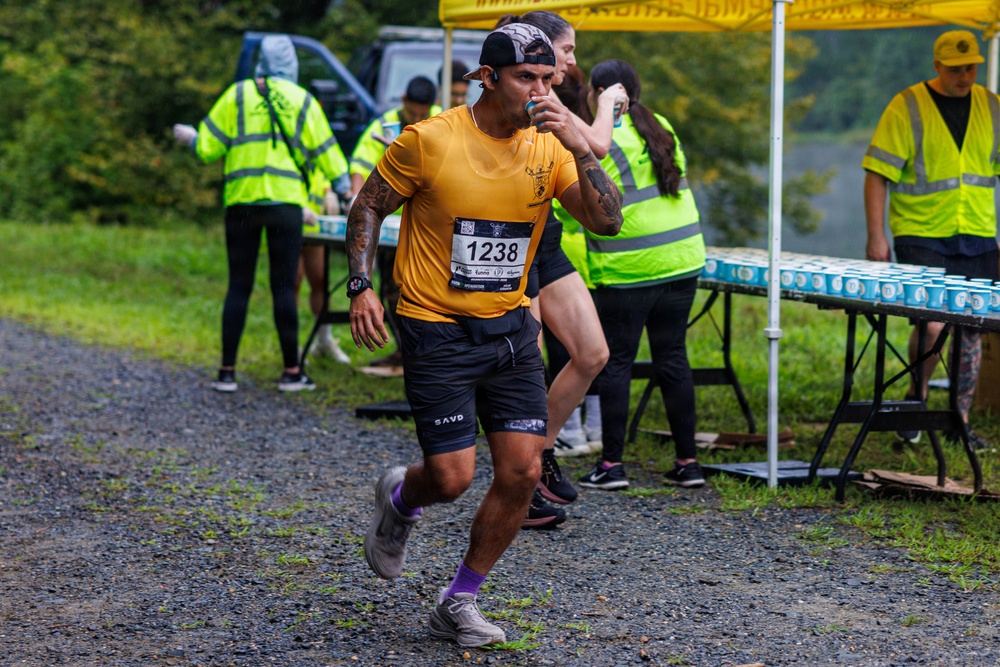 A male runner wearing bib number 1238 and a yellow shirt passes through a water/aid station during a road or trail race. He is drinking from a small cup while continuing to run on a gravel path. Volunteers in high-visibility yellow safety vests are staffing the aid station table, which is stocked with rows of pre-filled cups.