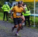 A male runner wearing bib number 1238 and a yellow shirt passes through a water/aid station during a road or trail race. He is drinking from a small cup while continuing to run on a gravel path. Volunteers in high-visibility yellow safety vests are staffing the aid station table, which is stocked with rows of pre-filled cups.