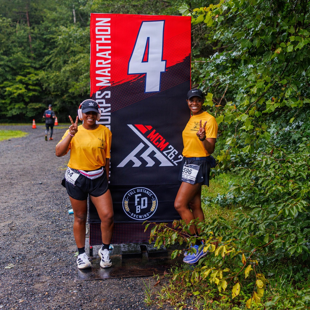 Two women in matching yellow shirts and black shorts pose with peace signs at mile marker 4 of the Marine Corps Marathon (MCM), a 26.2-mile race. Both wear race bibs (numbers 294 and 596) and black caps, smiling broadly at the camera. The setting is a gravel trail surrounded by green trees, with another runner visible in the background.