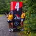 Two women in matching yellow shirts and black shorts pose with peace signs at mile marker 4 of the Marine Corps Marathon (MCM), a 26.2-mile race. Both wear race bibs (numbers 294 and 596) and black caps, smiling broadly at the camera. The setting is a gravel trail surrounded by green trees, with another runner visible in the background.