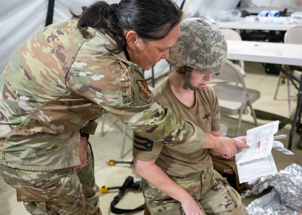 Two military personnel in camouflage uniforms are engaged in what appears to be a medical training or triage exercise inside a tent or temporary facility. One standing individual wearing OCP (Operational Camouflage Pattern) uniform with an Air Force patch is leaning over and pointing at a document or card held by a seated individual wearing ACU (Army Combat Uniform) pattern helmet and tan t-shirt. A mylar/emergency blanket is visible near the seated person, suggesting a casualty care scenario.