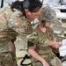 Two military personnel in camouflage uniforms are engaged in what appears to be a medical training or triage exercise inside a tent or temporary facility. One standing individual wearing OCP (Operational Camouflage Pattern) uniform with an Air Force patch is leaning over and pointing at a document or card held by a seated individual wearing ACU (Army Combat Uniform) pattern helmet and tan t-shirt. A mylar/emergency blanket is visible near the seated person, suggesting a casualty care scenario.