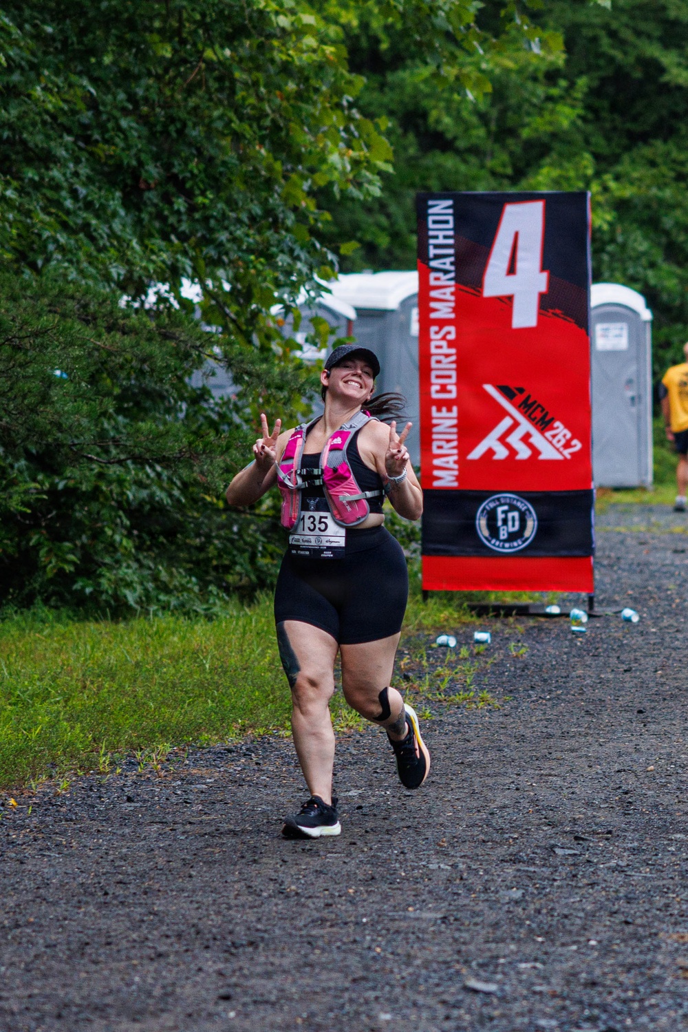 A female runner wearing bib number 135 is captured mid-stride at mile marker 4 of the Marine Corps Marathon (MCM 26.2). She is smiling broadly and making peace signs with both hands, displaying an enthusiastic and joyful demeanor. The race takes place on a gravel path surrounded by green trees, with portable toilets and scattered cups visible in the background.