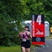 A female runner wearing bib number 135 is captured mid-stride at mile marker 4 of the Marine Corps Marathon (MCM 26.2). She is smiling broadly and making peace signs with both hands, displaying an enthusiastic and joyful demeanor. The race takes place on a gravel path surrounded by green trees, with portable toilets and scattered cups visible in the background.