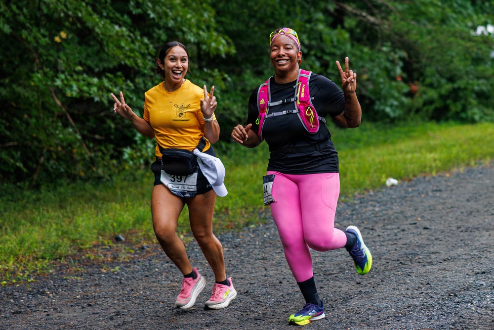 Two women are running together on a gravel path during what appears to be a trail race or fun run. Both are smiling broadly and making peace/victory signs with their hands, conveying joy and camaraderie. They are wearing race bibs and athletic gear appropriate for running.