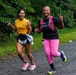 Two women are running together on a gravel path during what appears to be a trail race or fun run. Both are smiling broadly and making peace/victory signs with their hands, conveying joy and camaraderie. They are wearing race bibs and athletic gear appropriate for running.