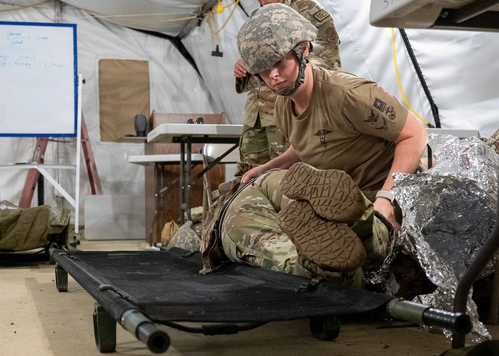 A U.S. Army soldier wearing ACU/OCP camouflage uniform and a combat helmet is kneeling beside a military field cot inside a tent structure, appearing to attend to a simulated casualty wrapped in a thermal/mylar emergency blanket. The setting appears to be a field medical training exercise, indicated by the whiteboard with notes, medical insignia on the soldier's uniform, and the tent environment. A second soldier is visible in the background.