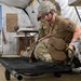 A U.S. Army soldier wearing ACU/OCP camouflage uniform and a combat helmet is kneeling beside a military field cot inside a tent structure, appearing to attend to a simulated casualty wrapped in a thermal/mylar emergency blanket. The setting appears to be a field medical training exercise, indicated by the whiteboard with notes, medical insignia on the soldier's uniform, and the tent environment. A second soldier is visible in the background.