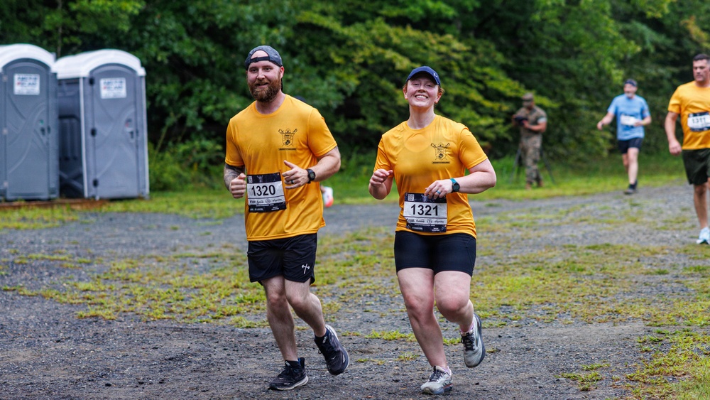 Two runners wearing matching yellow t-shirts and race bibs numbered 1320 and 1321 are running together on a gravel path during an organized race event. They appear to be in good spirits, with the woman smiling broadly at the camera. The setting is an outdoor trail race with portable toilets, lush green trees, and other participants visible in the background.