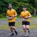 Two runners wearing matching yellow t-shirts and race bibs numbered 1320 and 1321 are running together on a gravel path during an organized race event. They appear to be in good spirits, with the woman smiling broadly at the camera. The setting is an outdoor trail race with portable toilets, lush green trees, and other participants visible in the background.