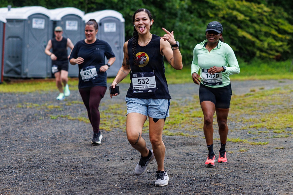 Three women are participating in an outdoor running race on a gravel path, with portable toilets and green trees visible in the background. The foreground runner, wearing bib number 532 and a black sleeveless shirt, is making a playful gesture with her tongue out and a hand sign toward the camera. The other runners wear bibs 424 and 1429, all dressed in athletic gear appropriate for a road or trail race.