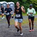 Three women are participating in an outdoor running race on a gravel path, with portable toilets and green trees visible in the background. The foreground runner, wearing bib number 532 and a black sleeveless shirt, is making a playful gesture with her tongue out and a hand sign toward the camera. The other runners wear bibs 424 and 1429, all dressed in athletic gear appropriate for a road or trail race.