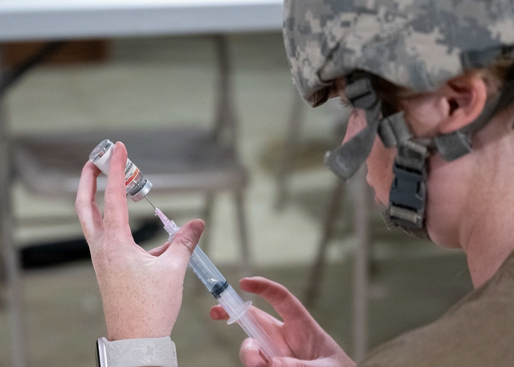 A person wearing U.S. military camouflage uniform and helmet is drawing medication from a small vial into a syringe. The individual appears to be a military medic or healthcare provider preparing an injection, likely in a field or temporary medical setting. Folding chairs and tables are visible in the blurred background.