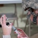 A person wearing U.S. military camouflage uniform and helmet is drawing medication from a small vial into a syringe. The individual appears to be a military medic or healthcare provider preparing an injection, likely in a field or temporary medical setting. Folding chairs and tables are visible in the blurred background.