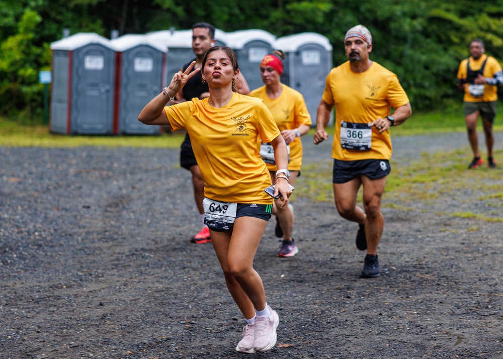 A group of runners participating in an organized road race on a gravel surface, all wearing matching yellow t-shirts. The foreground runner, bib number 649, is posing playfully for the camera with a peace sign and puckered lips while running. The background shows portable toilets, lush green trees, and additional runners including bib number 366.