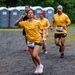 A group of runners participating in an organized road race on a gravel surface, all wearing matching yellow t-shirts. The foreground runner, bib number 649, is posing playfully for the camera with a peace sign and puckered lips while running. The background shows portable toilets, lush green trees, and additional runners including bib number 366.
