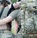 Two military personnel are shown from behind, with one appearing to support or assist the other. The individual on the right is wearing a large multicam tactical/medical backpack with visible scissors and red medical supplies. One person has a visibly injured or simulated-wound arm draped over the other's shoulder.