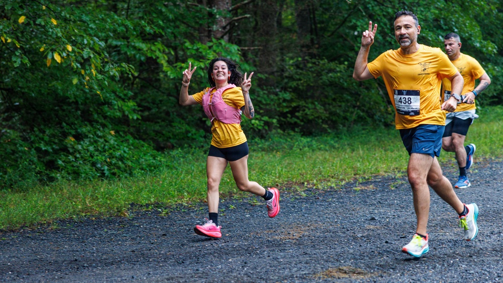 Three runners are participating in an organized road/trail race on a gravel path surrounded by dense green forest. The lead female runner is smiling and making peace signs with both hands, while a male runner wearing bib number 438 runs beside her, also making a peace sign. A third runner follows behind them, all wearing matching yellow team shirts.