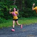 Three runners are participating in an organized road/trail race on a gravel path surrounded by dense green forest. The lead female runner is smiling and making peace signs with both hands, while a male runner wearing bib number 438 runs beside her, also making a peace sign. A third runner follows behind them, all wearing matching yellow team shirts.