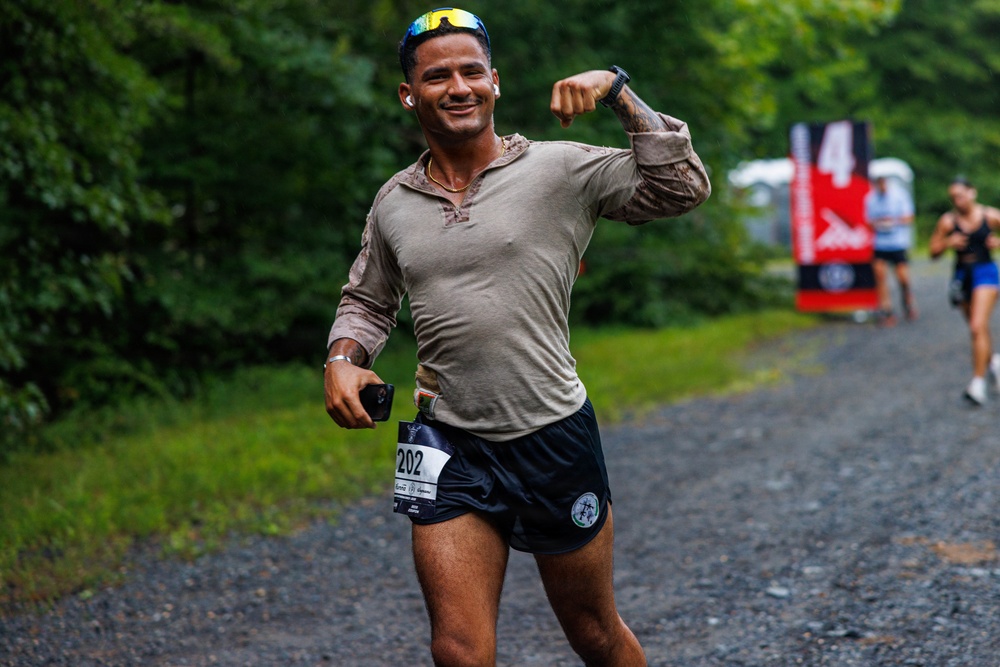 A male runner wearing bib number 202 is captured mid-race on a gravel path surrounded by green trees, smiling and flexing his right arm toward the camera. He is dressed in a long-sleeve gray shirt and dark shorts, with sunglasses pushed up on his head and wireless earbuds in. Other runners and a race marker banner are visible in the background.