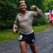A male runner wearing bib number 202 is captured mid-race on a gravel path surrounded by green trees, smiling and flexing his right arm toward the camera. He is dressed in a long-sleeve gray shirt and dark shorts, with sunglasses pushed up on his head and wireless earbuds in. Other runners and a race marker banner are visible in the background.