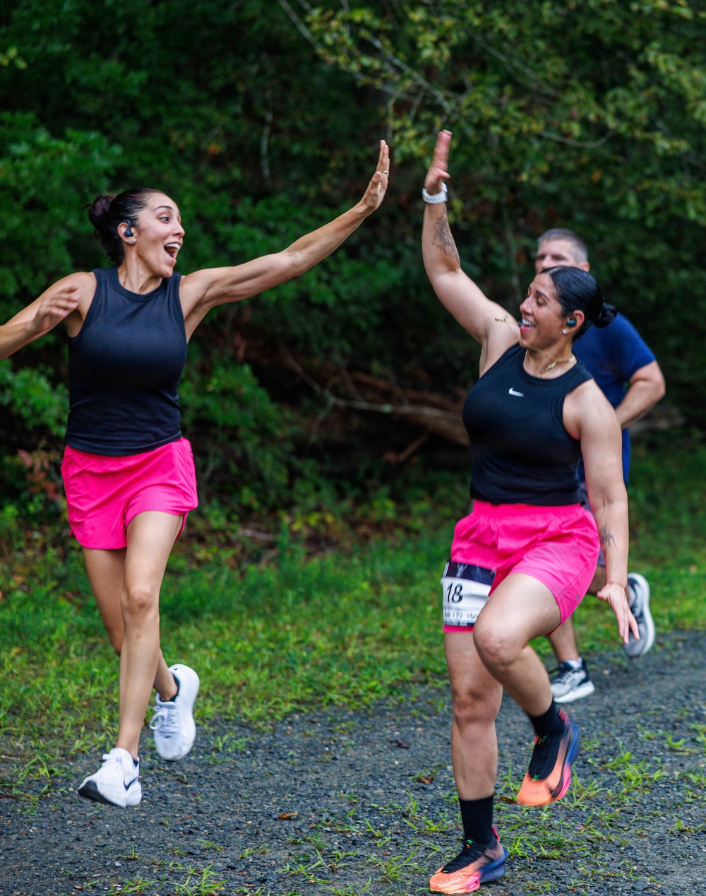Two women in matching black tank tops and bright pink shorts are high-fiving each other mid-run during what appears to be a road or trail race. Both are wearing wireless earbuds and the runner on the right displays a race bib numbered '18'. A male runner in a blue shirt is visible in the background.