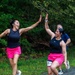 Two women in matching black tank tops and bright pink shorts are high-fiving each other mid-run during what appears to be a road or trail race. Both are wearing wireless earbuds and the runner on the right displays a race bib numbered '18'. A male runner in a blue shirt is visible in the background.