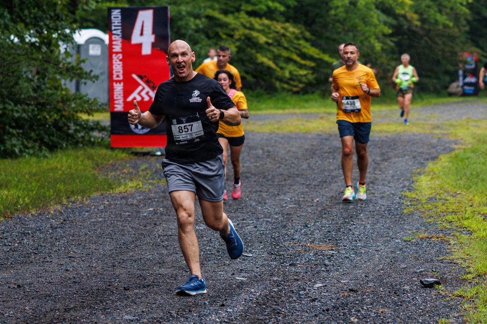 A group of runners competing in the Marine Corps Marathon event on a gravel trail path surrounded by trees. The lead runner, bib number 857, is a bald man in a black shirt and gray shorts giving two thumbs up with an enthusiastic expression. Several other runners follow behind, many wearing matching yellow shirts, with race signage visible in the background.