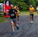 A group of runners competing in the Marine Corps Marathon event on a gravel trail path surrounded by trees. The lead runner, bib number 857, is a bald man in a black shirt and gray shorts giving two thumbs up with an enthusiastic expression. Several other runners follow behind, many wearing matching yellow shirts, with race signage visible in the background.