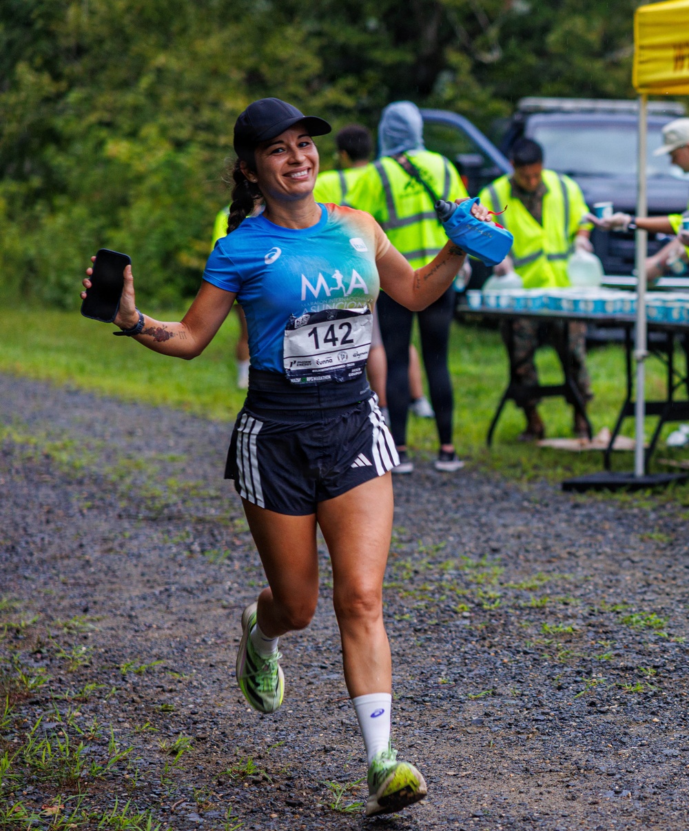 A female runner wearing bib number 142 is captured mid-stride on a gravel path during what appears to be an organized road or trail race. She is smiling broadly, holding a smartphone in her left hand and a hydration flask in her right, wearing an ASICS blue shirt branded 'MIA' and Adidas black shorts. In the background, race volunteers in high-visibility yellow vests are stationed at a water/aid station table.
