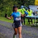 A female runner wearing bib number 142 is captured mid-stride on a gravel path during what appears to be an organized road or trail race. She is smiling broadly, holding a smartphone in her left hand and a hydration flask in her right, wearing an ASICS blue shirt branded 'MIA' and Adidas black shorts. In the background, race volunteers in high-visibility yellow vests are stationed at a water/aid station table.