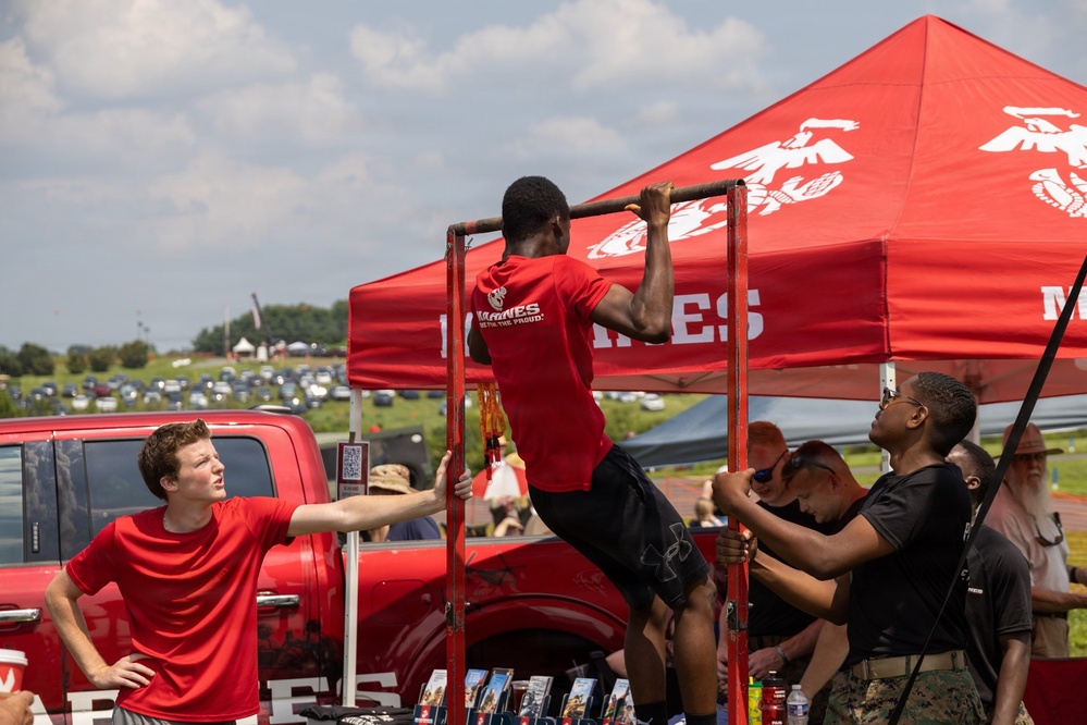 A U.S. Marines recruiting booth is set up at an outdoor public event, featuring a pull-up bar challenge. A young man in a red Marines t-shirt is performing pull-ups on a portable bar frame while others observe and assist. The booth is prominently branded with the Marines eagle, globe, and anchor insignia on a red canopy tent.