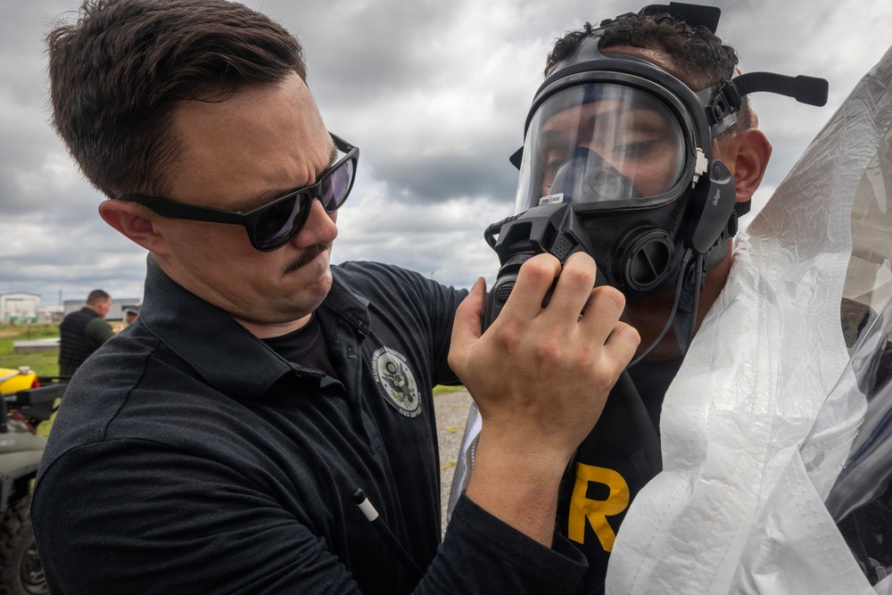 Two individuals are engaged in what appears to be a CBRN (Chemical, Biological, Radiological, Nuclear) training or preparedness exercise. One person wearing a black polo shirt with an official emblem and sunglasses is assisting another person in fitting or adjusting a full-face gas mask. The second individual is also wearing a white protective suit, suggesting a hazmat or emergency response training scenario.