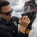 Two individuals are engaged in what appears to be a CBRN (Chemical, Biological, Radiological, Nuclear) training or preparedness exercise. One person wearing a black polo shirt with an official emblem and sunglasses is assisting another person in fitting or adjusting a full-face gas mask. The second individual is also wearing a white protective suit, suggesting a hazmat or emergency response training scenario.