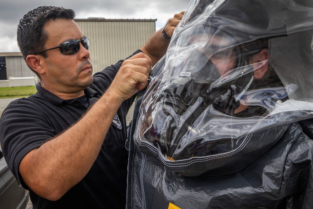 A man wearing sunglasses and a dark polo shirt is working with a large transparent plastic enclosure or protective covering, while another person is visible inside or behind the clear material wearing a helmet. The setting appears to be an outdoor area near an industrial or aviation facility. The activity suggests preparation or assistance with specialized protective equipment.