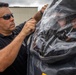 A man wearing sunglasses and a dark polo shirt is working with a large transparent plastic enclosure or protective covering, while another person is visible inside or behind the clear material wearing a helmet. The setting appears to be an outdoor area near an industrial or aviation facility. The activity suggests preparation or assistance with specialized protective equipment.