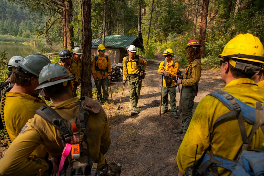 A group of wildland firefighters in standard Nomex fire-resistant gear are gathered on a dirt path in a forested area near a body of water, appearing to conduct a briefing or debriefing. The crew members are equipped with hand tools such as Pulaskis and wearing hard hats of various colors including yellow, white, gray, and pink. A small structure with a green metal roof is visible in the background among the trees.