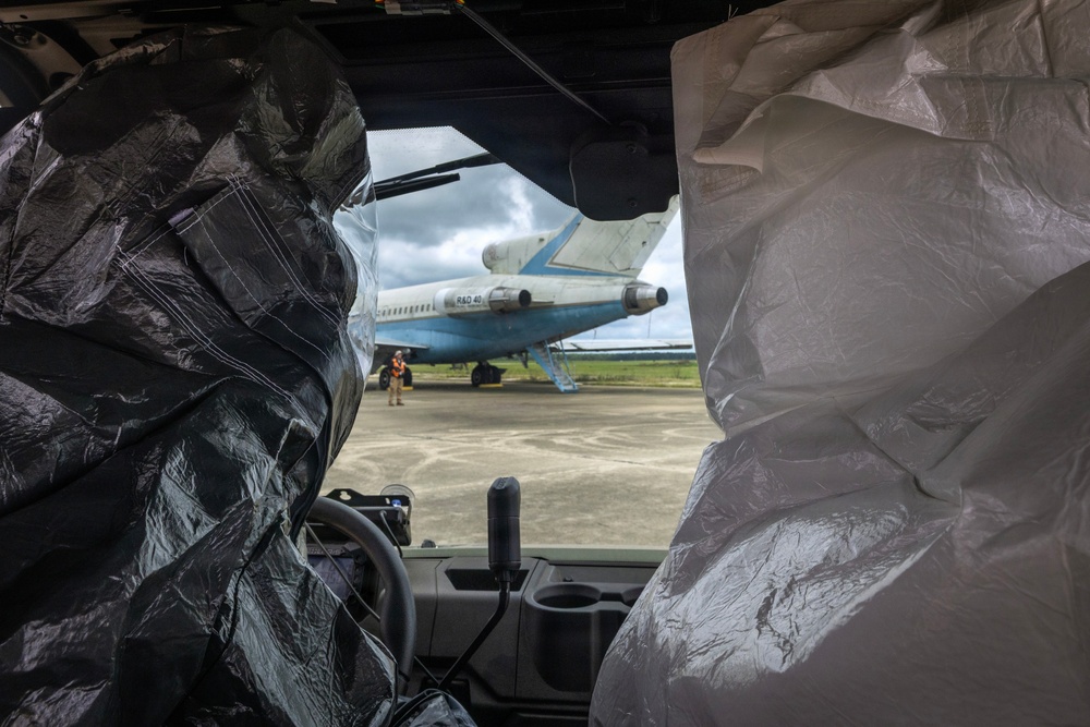 The image is taken from inside a vehicle, looking through the windshield at a parked commercial aircraft on an airport tarmac. The vehicle's seats are covered with black and silver/gray plastic protective covers. At least one ground crew member in a high-visibility vest is visible near the aircraft.