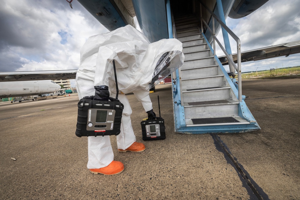 A person wearing a full white hazmat/Tyvek protective suit with an enclosed helmet visor and orange rubber boots is conducting what appears to be a hazardous materials inspection or detection operation at an airport tarmac. The individual is bent over near the base of an aircraft boarding stairway, carrying two black portable gas detection or multi-gas monitoring devices. The setting is an outdoor airport apron with other aircraft visible in the background under a partly cloudy sky.