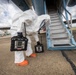A person wearing a full white hazmat/Tyvek protective suit with an enclosed helmet visor and orange rubber boots is conducting what appears to be a hazardous materials inspection or detection operation at an airport tarmac. The individual is bent over near the base of an aircraft boarding stairway, carrying two black portable gas detection or multi-gas monitoring devices. The setting is an outdoor airport apron with other aircraft visible in the background under a partly cloudy sky.