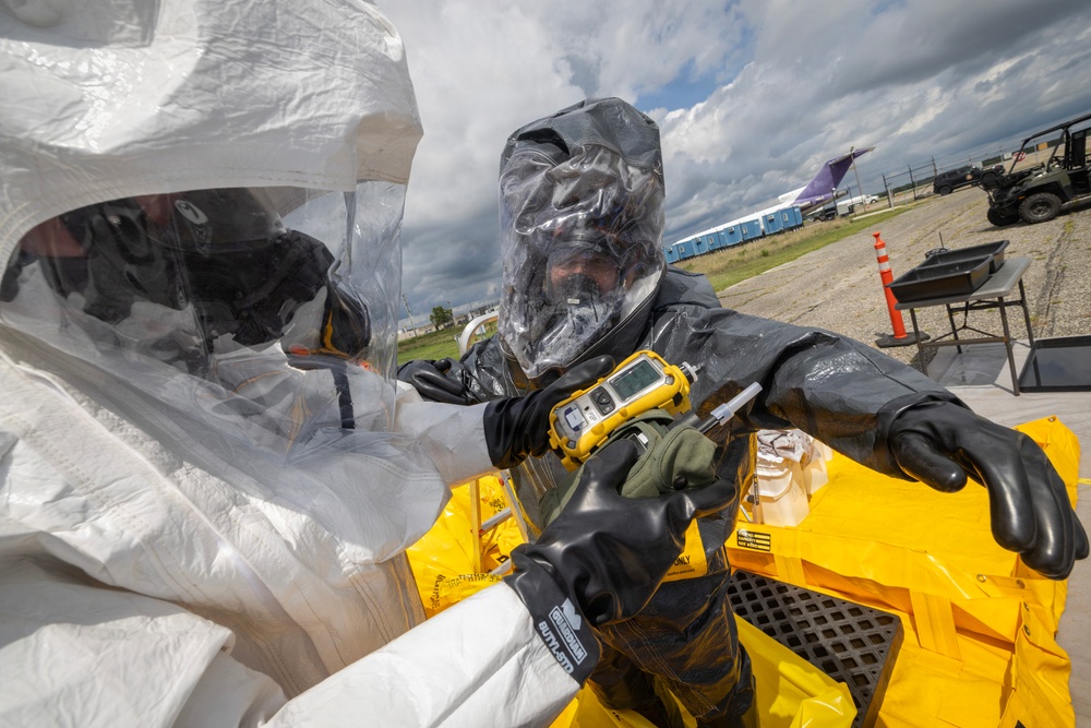 Two personnel in full hazmat/CBRN protective suits are conducting what appears to be a decontamination or hazardous materials response exercise at an airfield or military installation. One individual in a dark suit is holding a yellow multi-gas detector instrument, while the other wears a white protective suit. The setting includes aircraft, military vehicles, and portable infrastructure visible in the background.