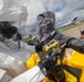 Two personnel in full hazmat/CBRN protective suits are conducting what appears to be a decontamination or hazardous materials response exercise at an airfield or military installation. One individual in a dark suit is holding a yellow multi-gas detector instrument, while the other wears a white protective suit. The setting includes aircraft, military vehicles, and portable infrastructure visible in the background.