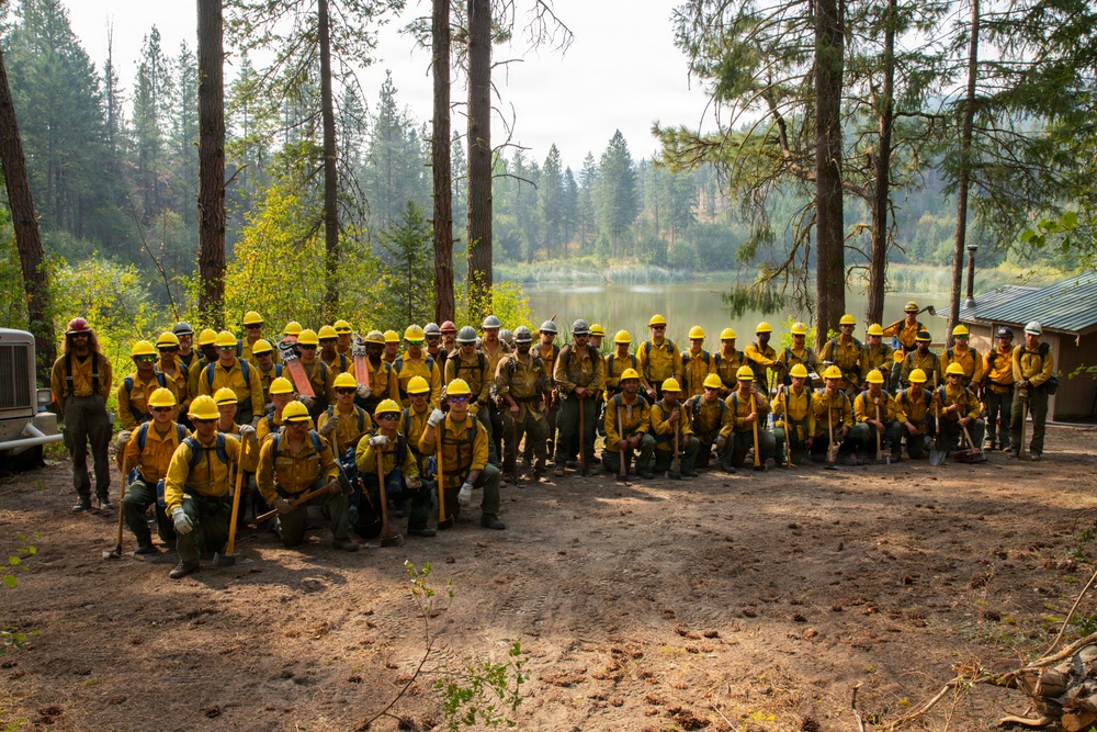 A large group of wildland firefighters poses for a group photograph in a forested area near a lake or river. The crew members are uniformly dressed in yellow Nomex fire-resistant shirts and yellow hard hats, carrying hand tools typical of wildland firefighting operations. The setting appears to be a staging or base camp area surrounded by tall conifers with hazy, smoke-affected skies visible in the background.