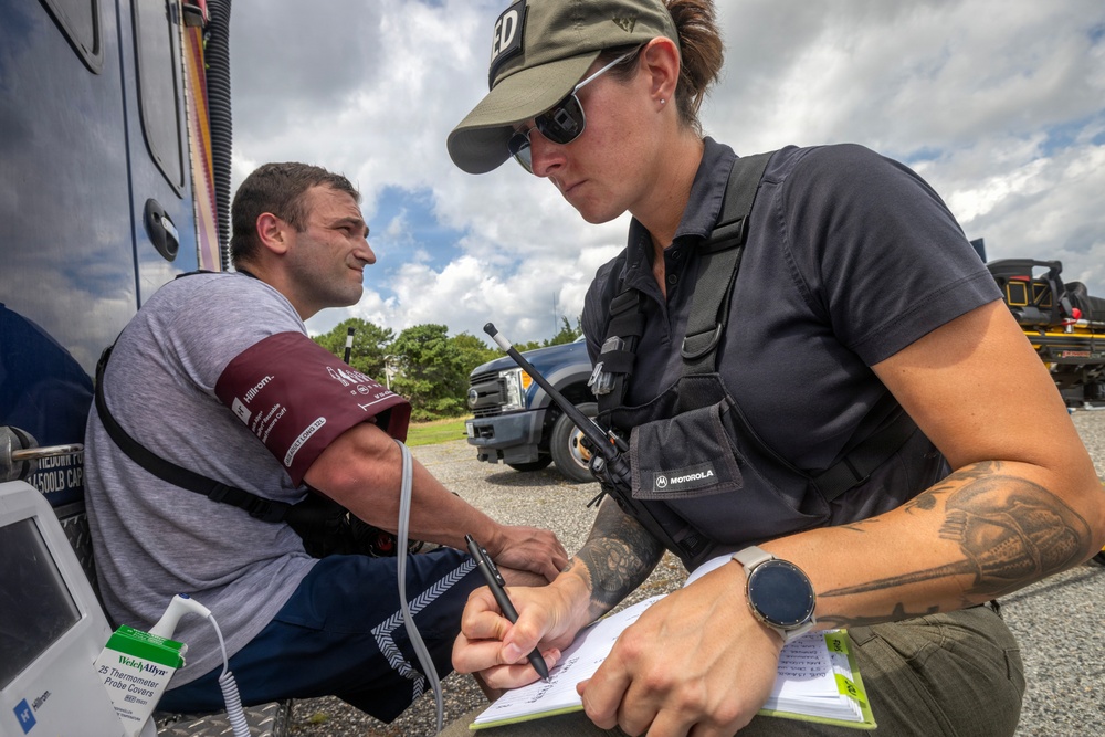 A female emergency medical or law enforcement responder is taking a male patient's blood pressure and recording notes outdoors near emergency vehicles. The responder is wearing tactical gear including a vest with a Motorola radio and a cap partially reading 'ED', suggesting an emergency or federal role. The scene appears to be a field medical assessment or triage situation.