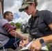 A female emergency medical or law enforcement responder is taking a male patient's blood pressure and recording notes outdoors near emergency vehicles. The responder is wearing tactical gear including a vest with a Motorola radio and a cap partially reading 'ED', suggesting an emergency or federal role. The scene appears to be a field medical assessment or triage situation.